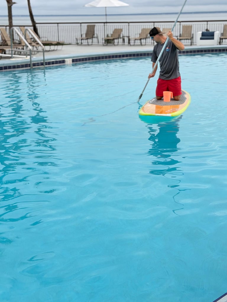 Man paddleboarding in a resort swimming pool using a paddle, wearing red shorts and gray shirt