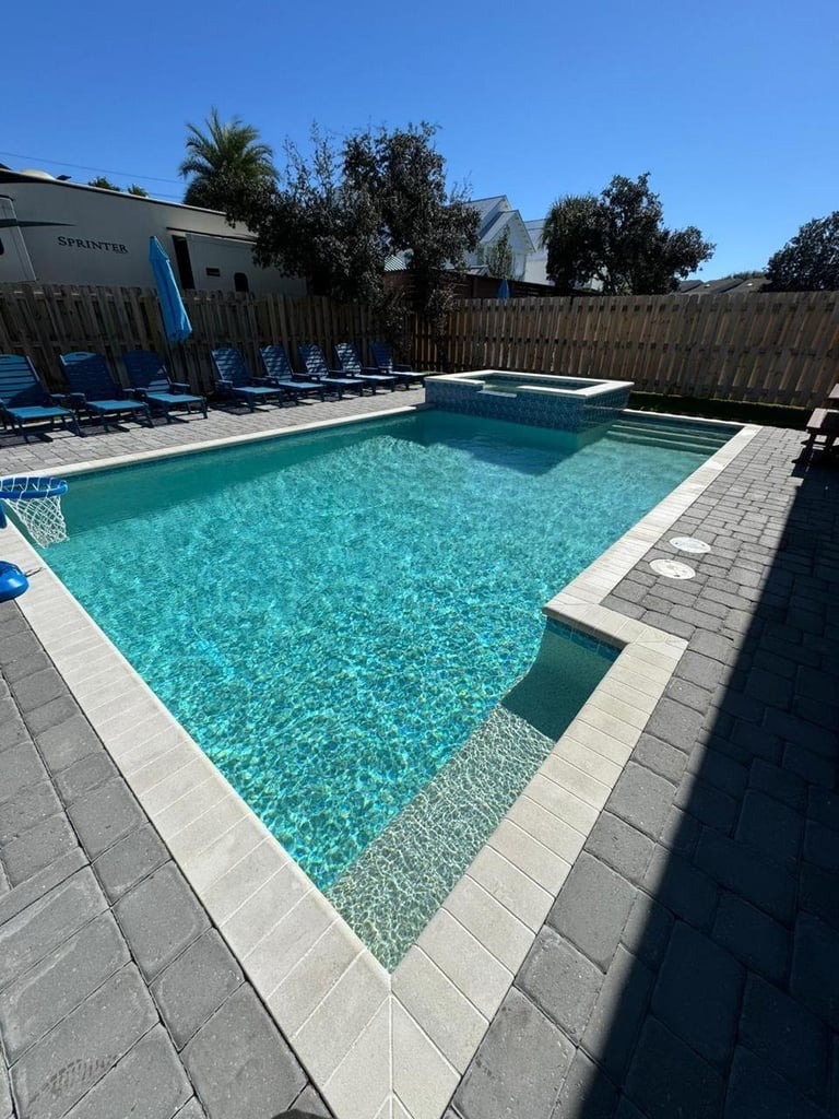 A bright turquoise swimming pool surrounded by gray pavers, lounge chairs, and a wooden fence on a sunny day