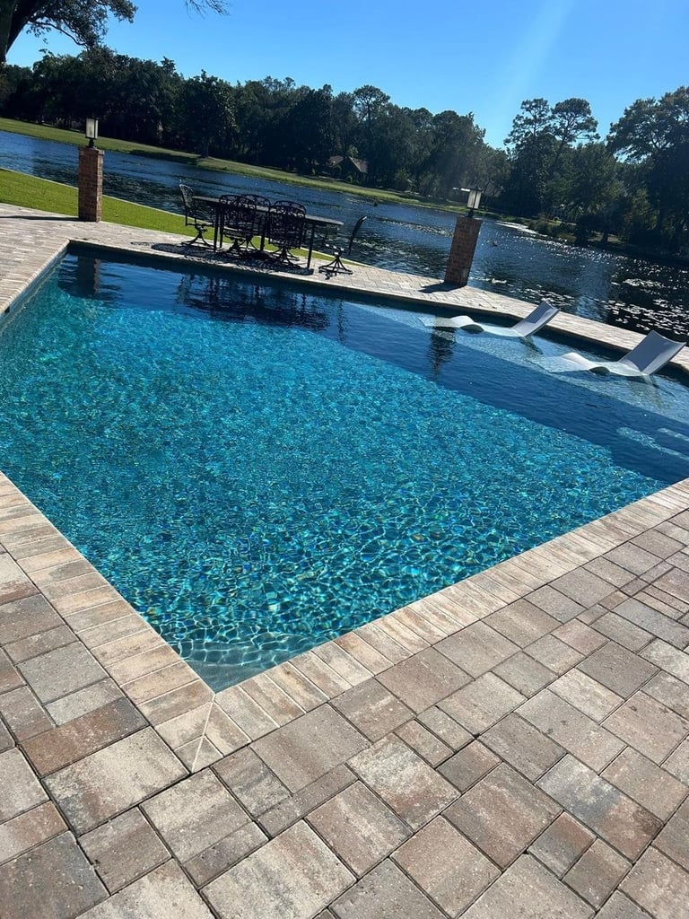 Crystal clear backyard swimming pool with paved deck overlooking a river and green lawns with trees