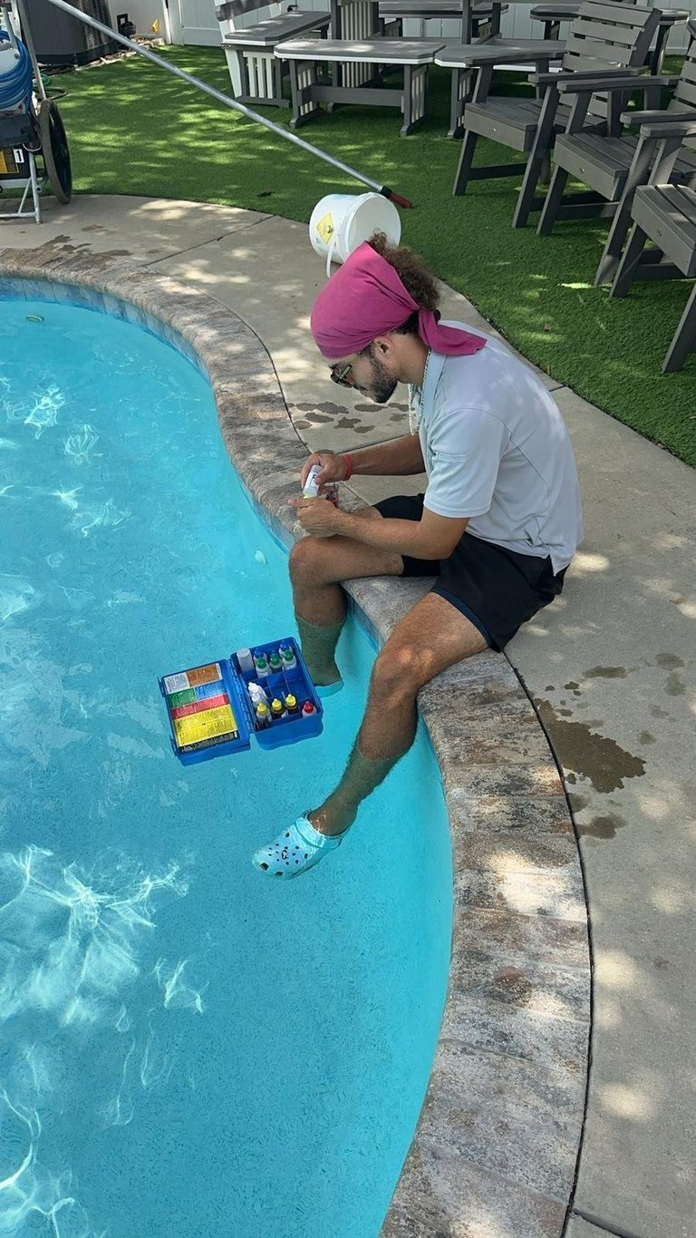 Person testing pool water chemistry using test kit while seated on pool deck with outdoor furniture visible in background